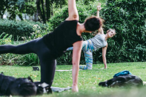 Zwei Personen praktizieren eine balancierende Yoga‑Pose auf einer Wiese, umgeben von üppigem Grün.
