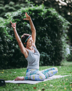 Person sitzt im Schneidersitz auf einer Yogamatte im Grünen und streckt beide Arme nach oben.