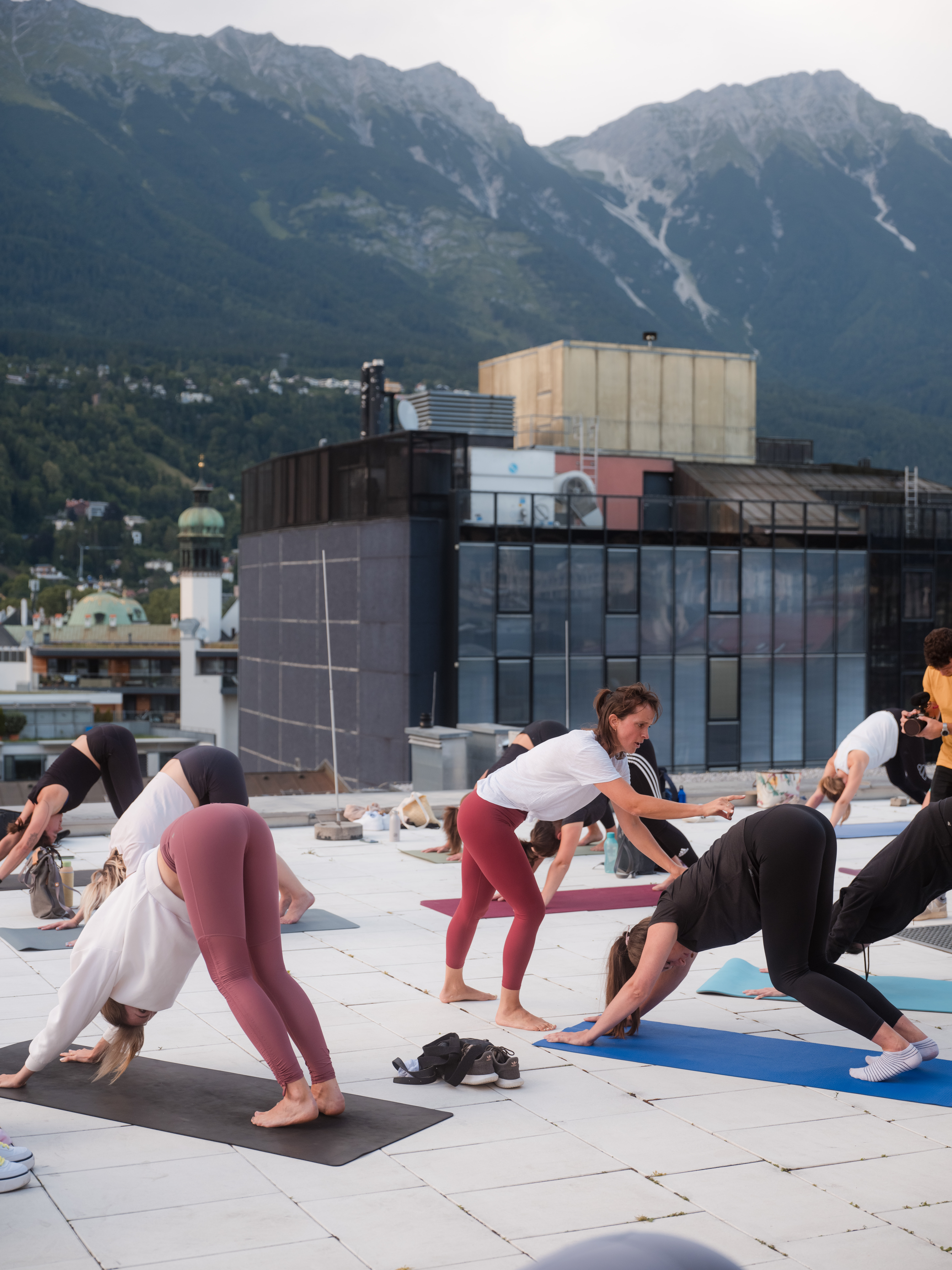 Personen auf einer Dachterrasse strecken die Arme nach oben während einer Yoga‑Übung, mit klarer Sicht auf die umliegenden Berge.