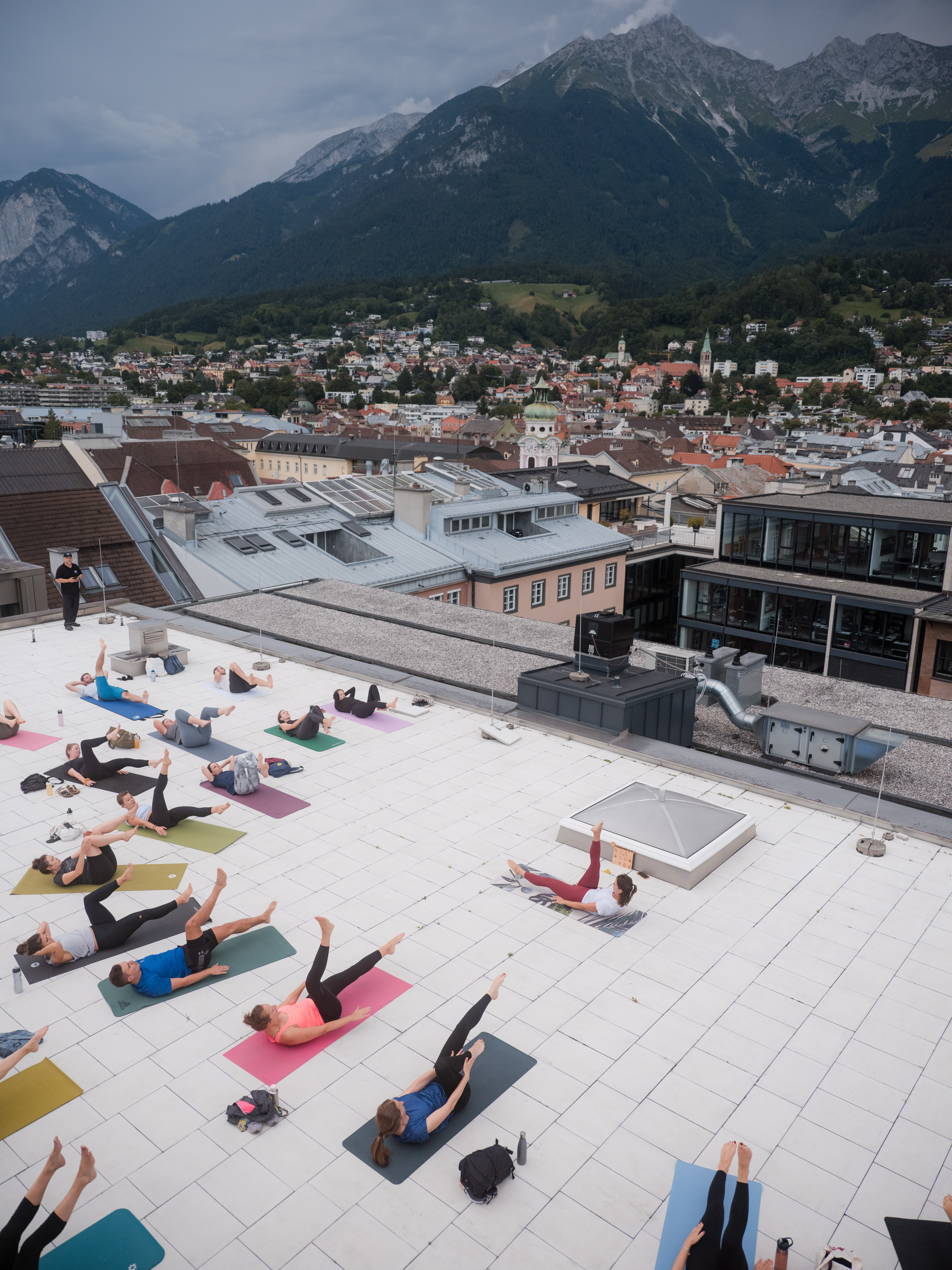 Viele Menschen üben gemeinsam Yoga auf einer Dachterrasse, umgeben von Stadtgebäuden und Berglandschaft.
