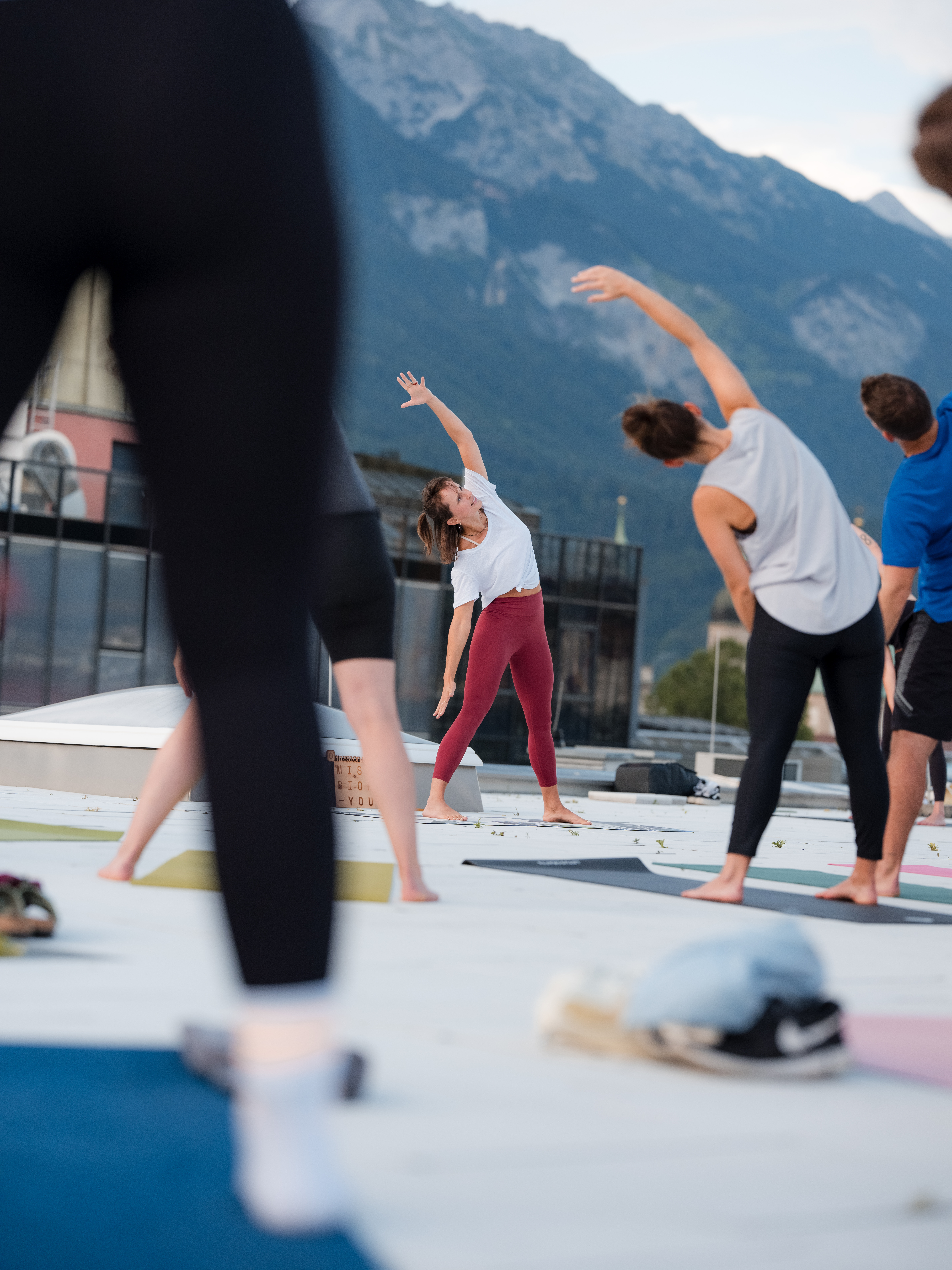 Yoga‑Gruppe auf einer Dachterrasse; Trainerin unterstützt eine Teilnehmerin bei einer Pose, im Hintergrund Stadt und Berge.